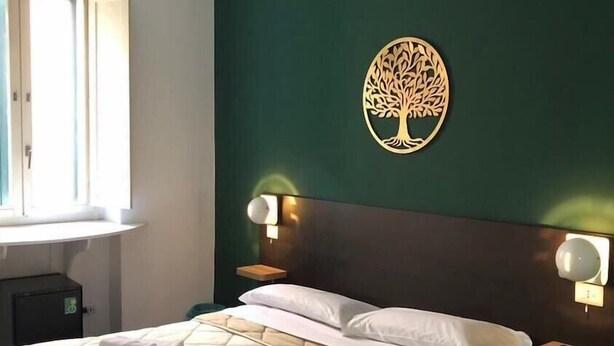 Een hotelkamer met een bed, twee bedlampen, een decoratieve boom aan de muur en een raam met gordijnen.
