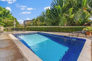 A rectangular swimming pool with clear blue water, surrounded by a concrete deck and a variety of tropical plants. - Cozy Island Escape - Near Waikiki & Diamond Head (Honolulu)