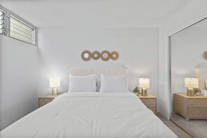 A bedroom with a large bed, two bedside tables with lamps, a wall-mounted mirror, and a window with blinds. - Cozy Island Escape - Near Waikiki & Diamond Head (Honolulu)