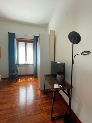 A room with a wooden floor, a desk with a computer, a standing lamp, and a window with curtains.