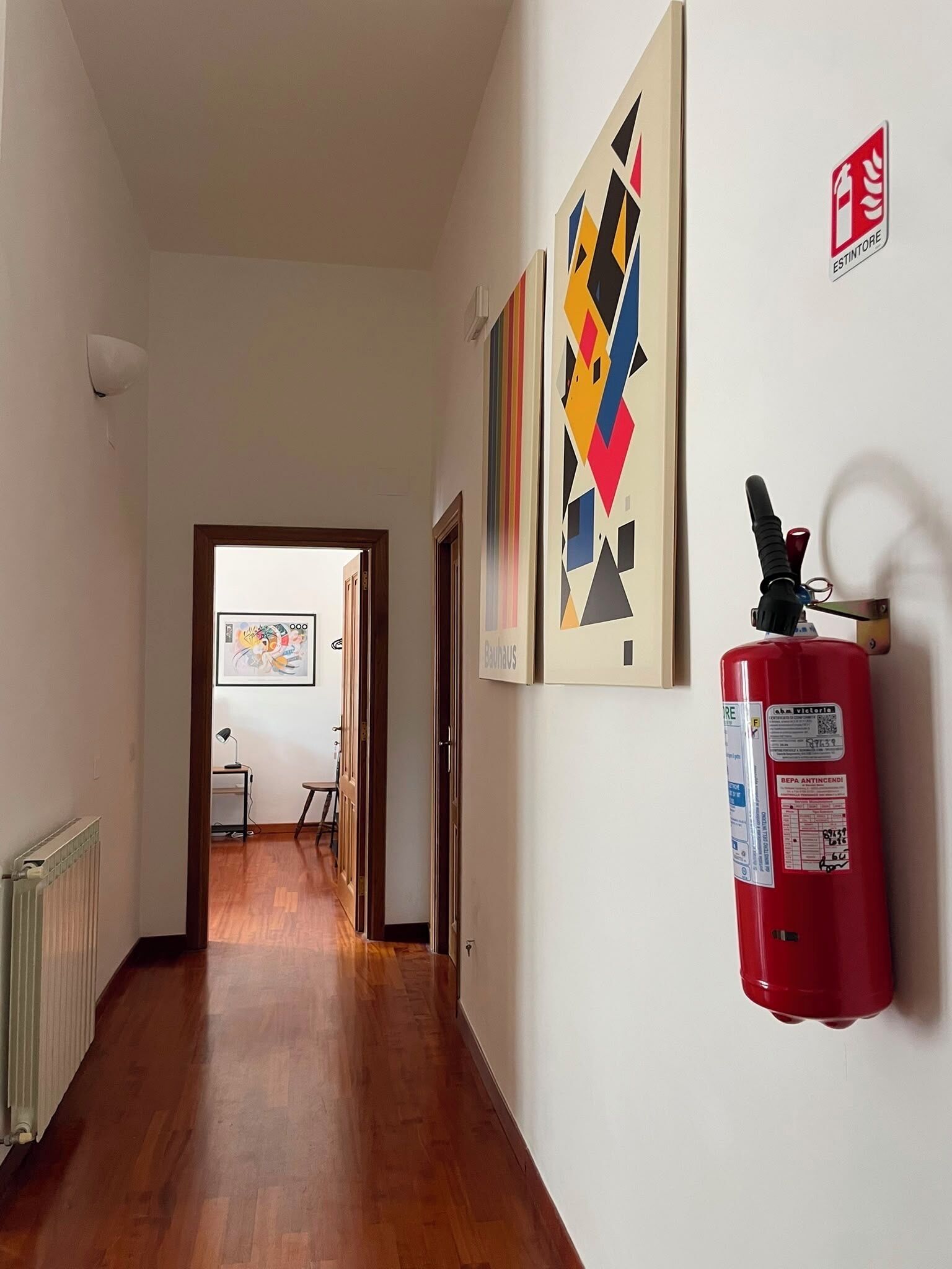 A hallway with wooden flooring, a fire extinguisher mounted on the wall, and framed artworks.