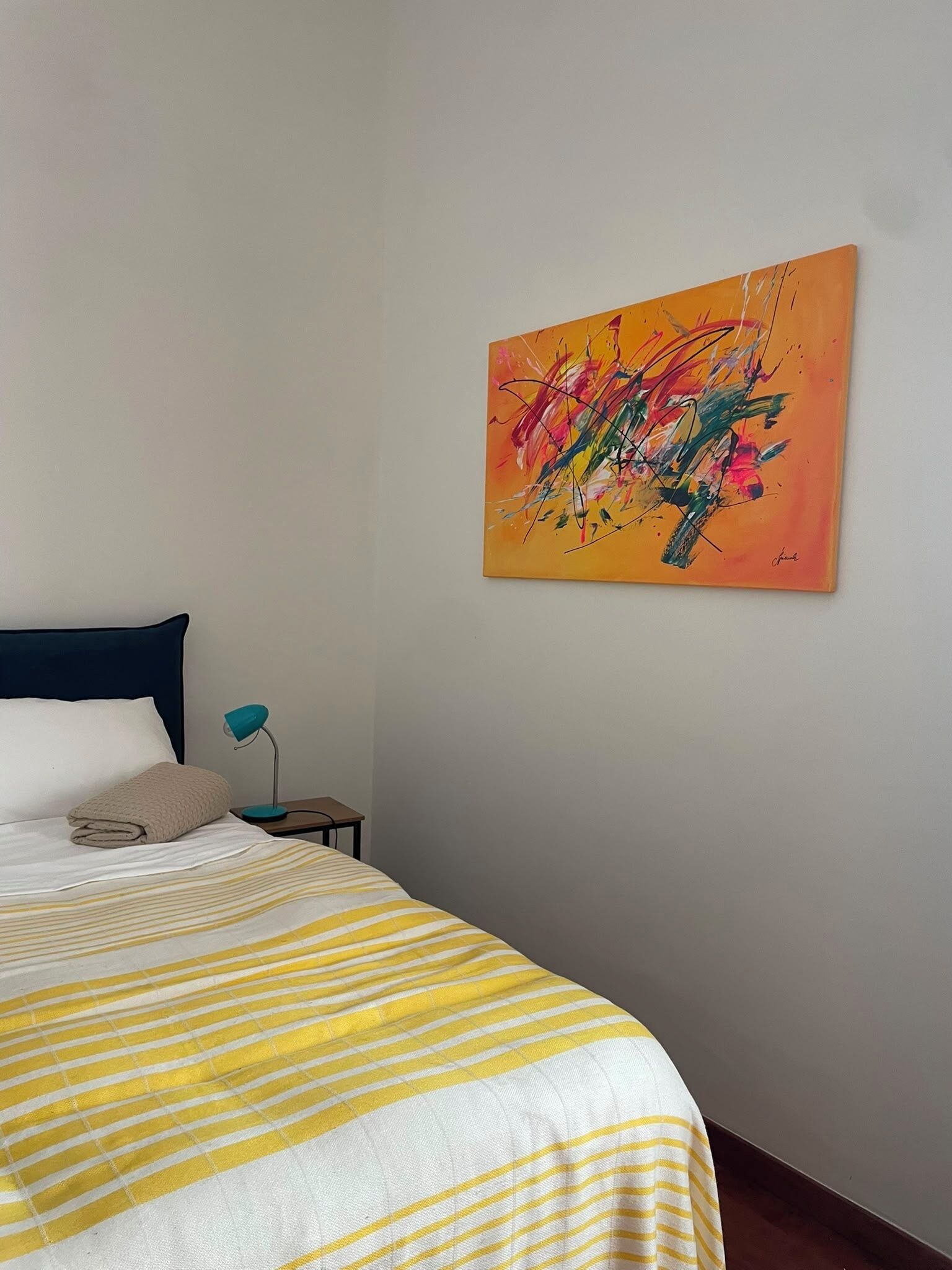 Ein Schlafzimmer mit einem Bett, einem Nachttisch, einer Lampe und einem farbenfrohen, abstrakten Gemälde an der Wand.