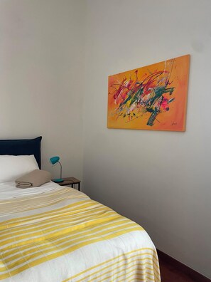 A bedroom with a bed, a nightstand, a lamp, and a colorful abstract painting on the wall. - Near Me:Mansio Portus,Civitavecchia, Railroad,Port, Cruise Ships,Rome,Etruscans (Civitavecchia)