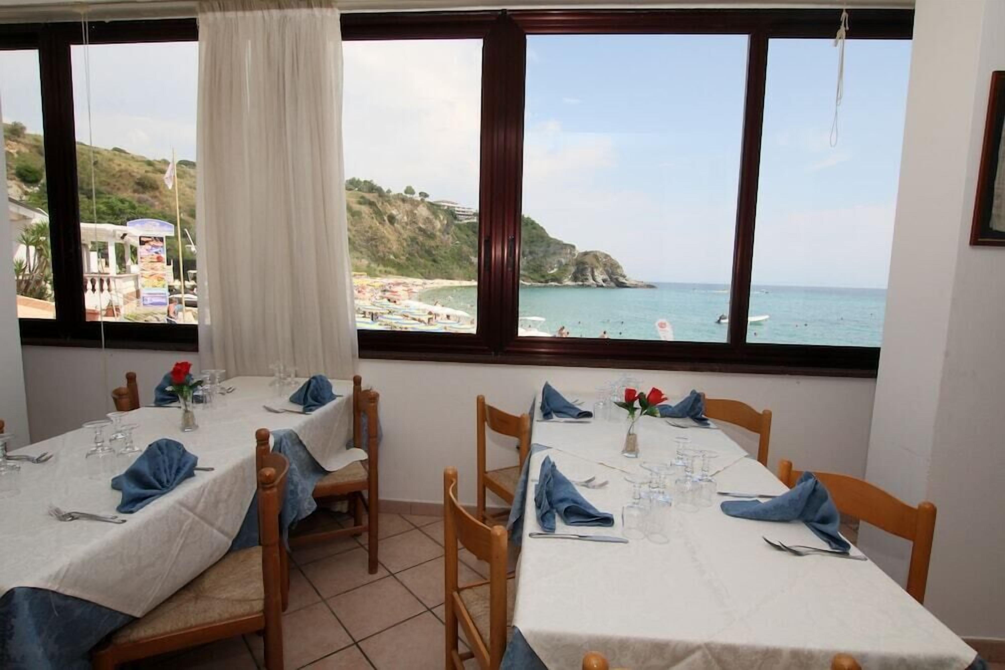A dining area with tables set for two, overlooking a beach with a rocky coastline.