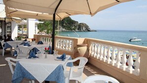 Un área de comedor al aire libre con mesas y sillas, vista al mar y un barco a lo lejos.