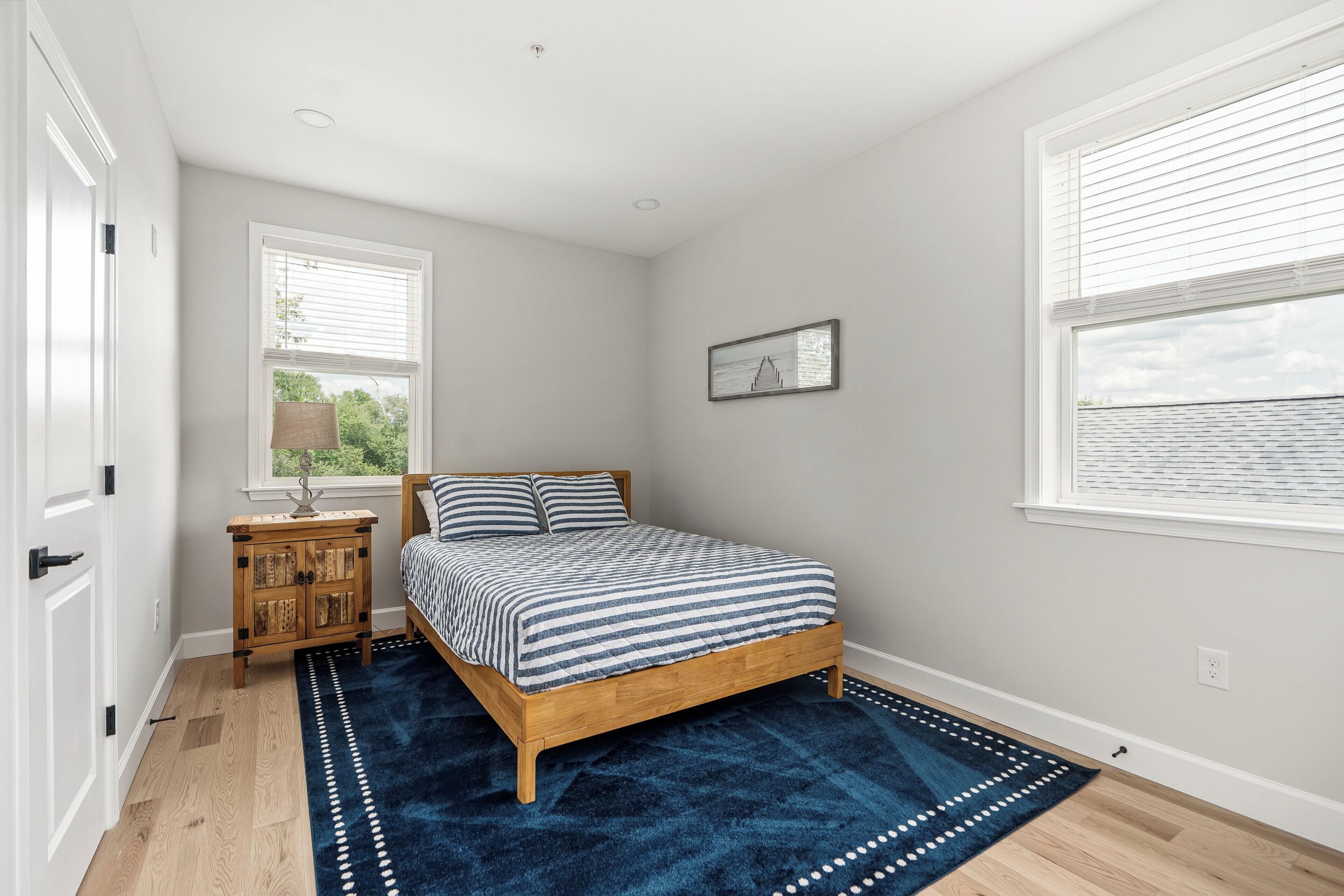 A bedroom with a wooden bed, a blue rug, a nightstand, a window with blinds, and a wall-mounted picture.