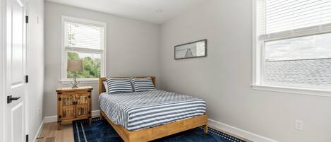 A bedroom with a wooden bed, a blue rug, a nightstand, a window with blinds, and a wall-mounted picture.