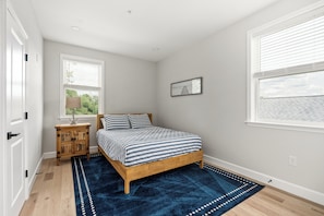 A bedroom with a wooden bed, a blue rug, a nightstand, a window with blinds, and a wall-mounted picture. - Stay Blue Heron, Coastal Comfort in Charlestown (Charlestown)