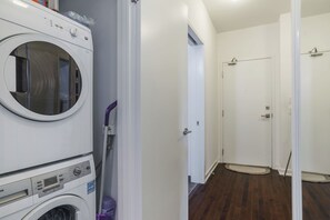 Une buanderie avec des machines à laver et des sèche-linge superposés, une porte blanche donnant sur un couloir et un plancher en bois.