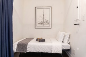 A single bed with a gray blanket and a rolled towel, a framed cityscape artwork on the wall, and a blue curtain partially visible. - CN Tower View 2-Bed+den, with parking 8 Guests (Toronto)
