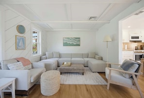 A modern living room with a sectional sofa, a small coffee table, and a round ottoman. There is a painting on the wall and a window with a view of the outdoors. - Walk To The Beach, Rooftop Deck With Water Views (Bourne)