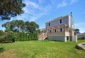 Une maison à deux étages avec une terrasse en bois, un escalier menant à l’étage supérieur et une cour clôturée.