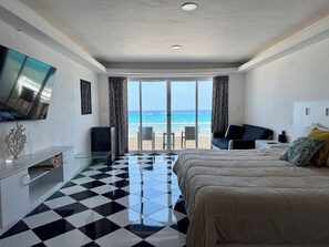 A hotel room with a checkered floor, a bed, a TV, a desk, and a view of the beach.