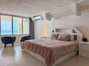 A bedroom with a large bed, a window with a view of the sea, and a modern air conditioning unit.