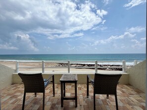 A patio with two wicker chairs and a table overlooking a beach with a rocky shoreline.