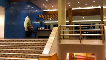 Een hotel lobby met een marmeren receptiebalie, een trap met houten leuning en een blauwe muur met een decoratieve spiegel.