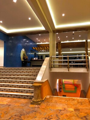 A hotel lobby with a marble reception desk, a staircase with a wooden railing, and a blue wall with a decorative mirror.