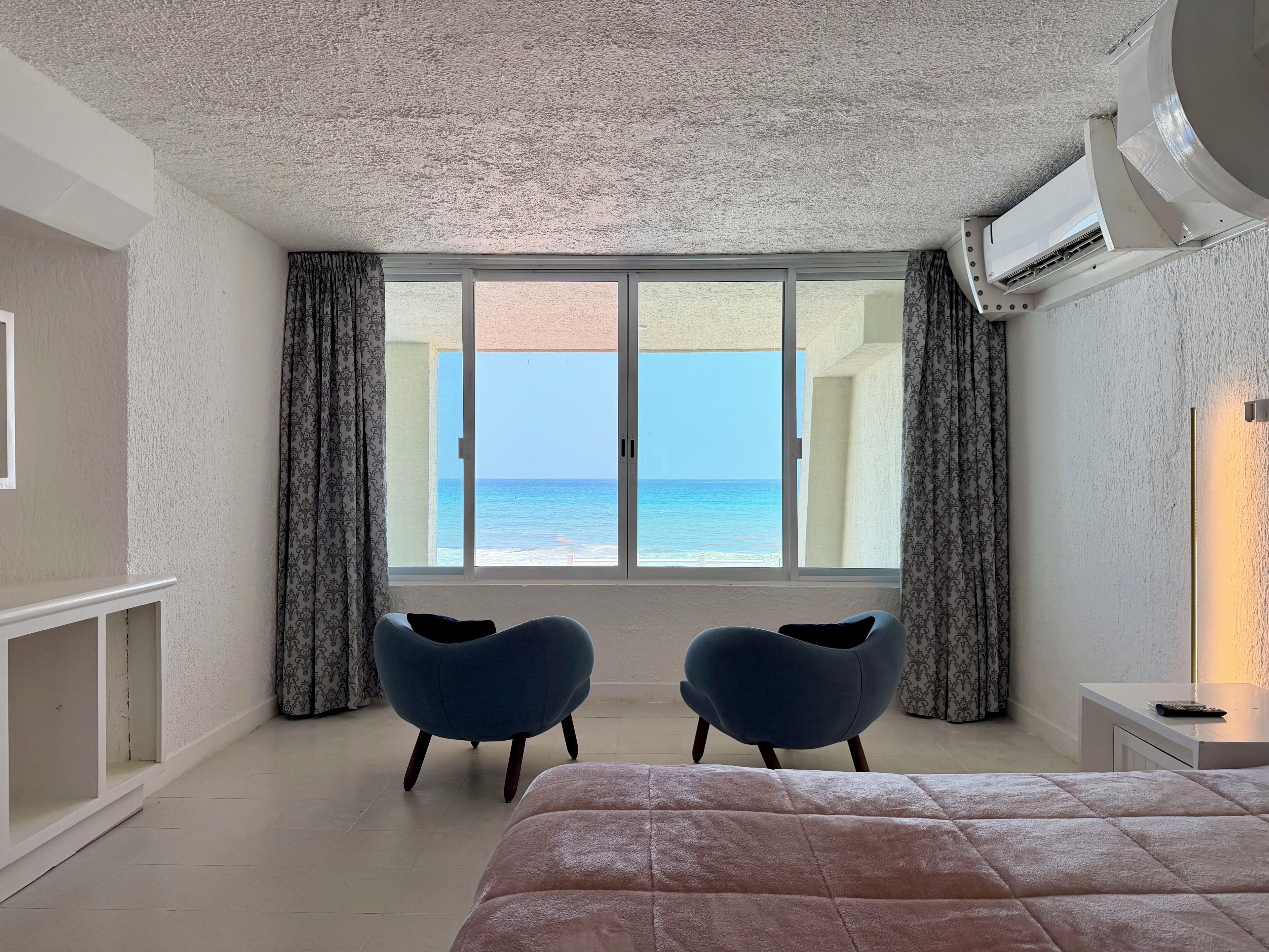 A bedroom with a large bed, two chairs, a TV, and a view of the sea.