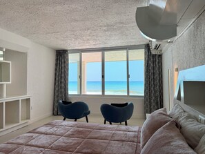 A bedroom with a large bed, two chairs, a TV, and a view of the sea through a large window.