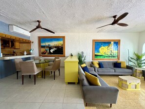 Un salon comprenant un coin repas, deux ventilateurs de plafond, un tableau et un canapé avec des coussins.