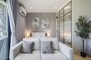 Ein modernes Schlafzimmer mit Schlafsofa, zwei Nachttischen mit Lampen, einem großen Fenster mit Jalousien und einer Zimmerpflanze.