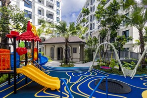 Ein Spielplatz mit Rutschen und Klettergerüsten, umgeben von Palmen und einem Gebäude mit Balkonen.