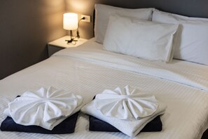 Ein ordentlich bezogenes Bett mit weißen Leinen, zwei auf dem Bett gefaltete Handtücher, ein weißes Kopfkissen und eine Nachttischlampe auf einem Nachttisch.
