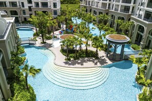 Una gran piscina al aire libre rodeada de palmeras y un pabellón, con un edificio de varios niveles al fondo.