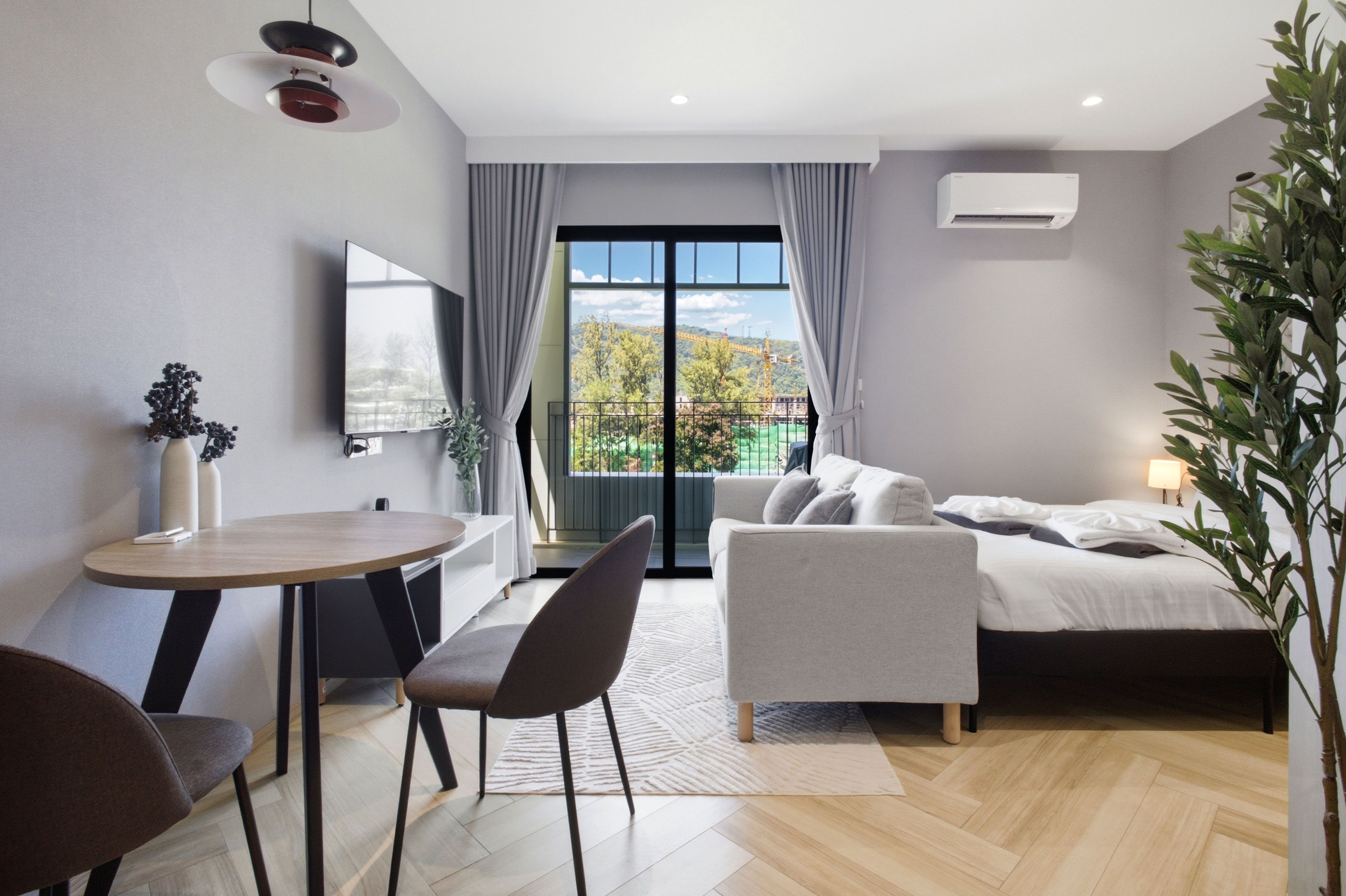Une chambre d’hôtel moderne équipée d’un lit, d’un canapé, d’une table à manger et d’une chaise. Une grande fenêtre offre une vue sur les arbres et un chantier de construction.