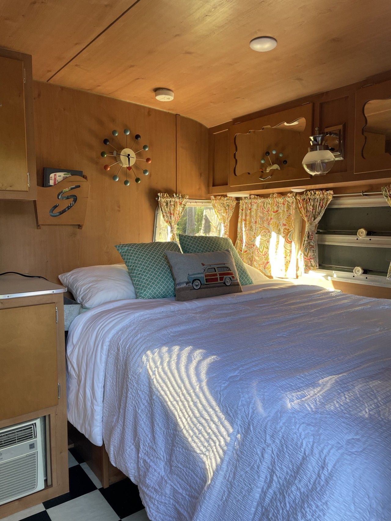 Un dormitorio de autocaravana acogedor con cama, almohadas, un reloj y una ventana con cortinas.