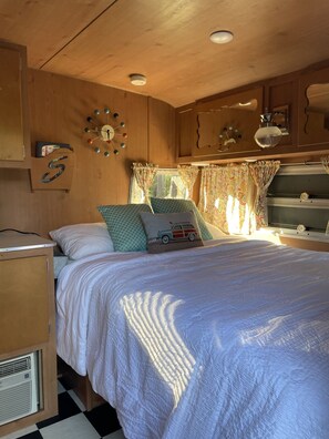 Un dormitorio de autocaravana acogedor con cama, almohadas, un reloj y una ventana con cortinas.