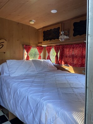 Habitación individual con una cama, techo de madera, ventana con cortinas rojas e iluminación fijada a la pared.