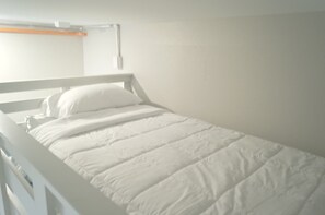 Una cama individual con cabecera y cubrecama blancos en una habitación con paredes lisas.