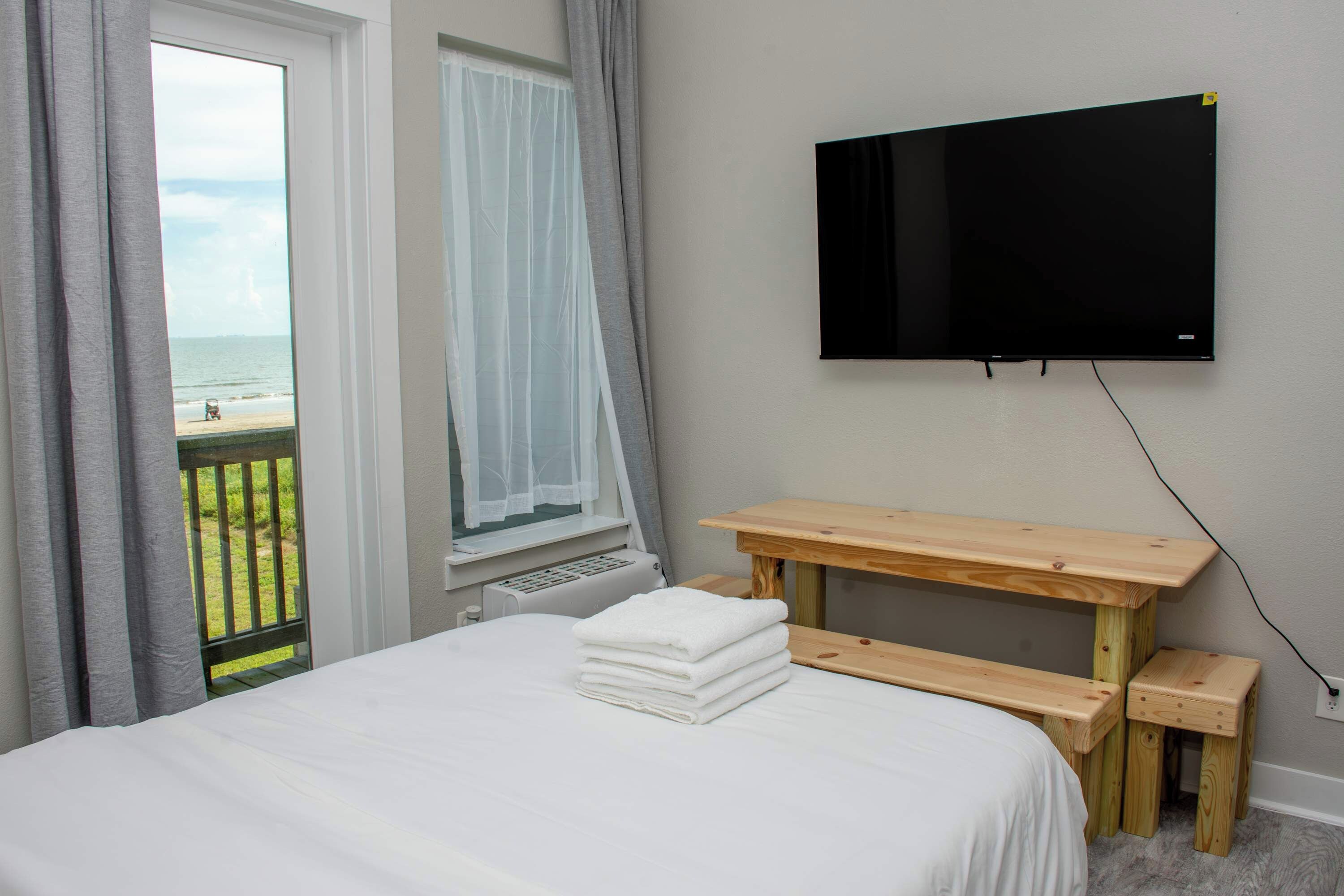 Une chambre à coucher avec un lit, une table en bois, un téléviseur fixé au mur et une fenêtre donnant sur la plage.