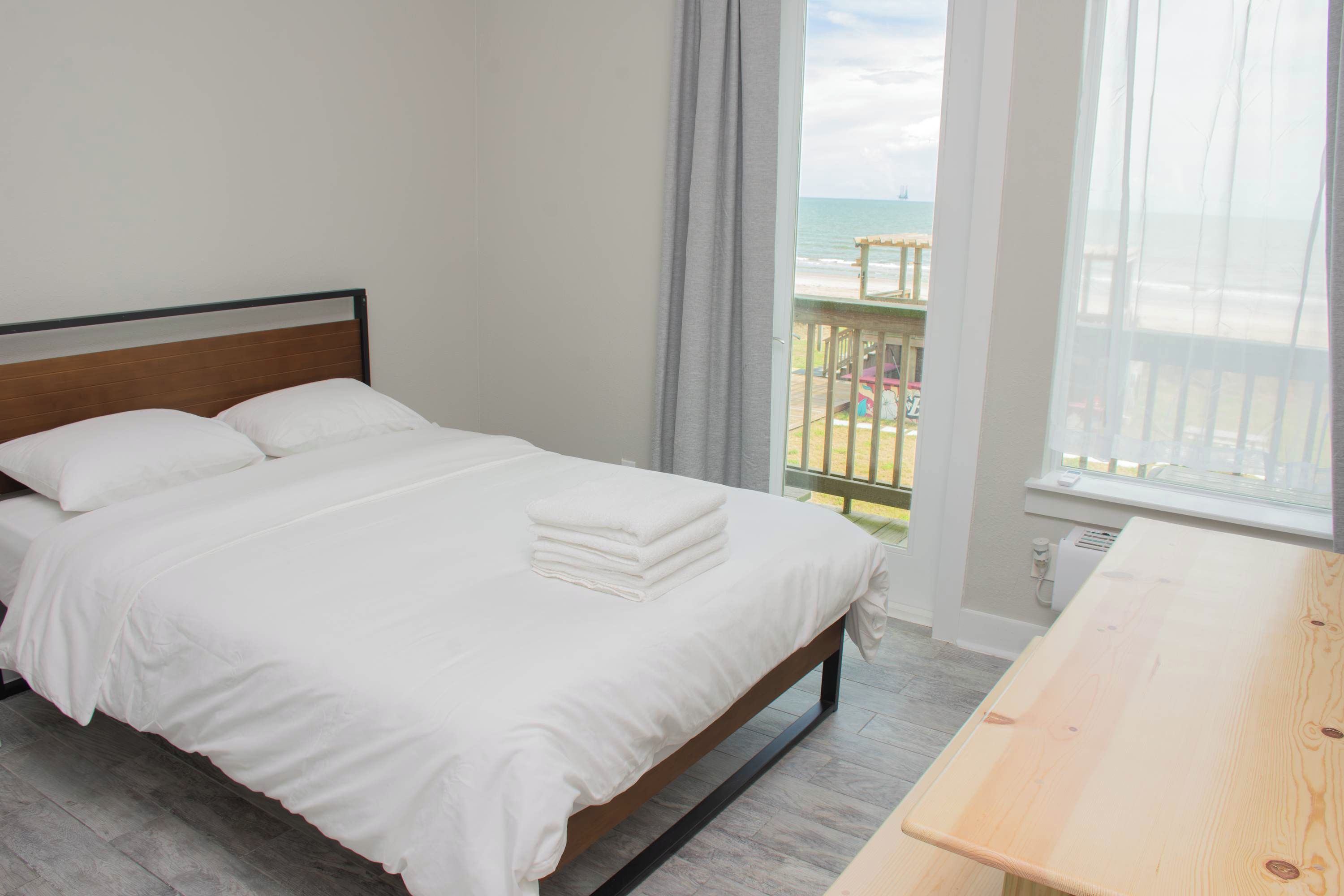 A hotel room with a bed, a wooden nightstand, and a view of the beach.