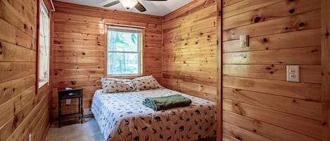 A wooden cabin room with a bed, a window, a ceiling fan, and a small table.