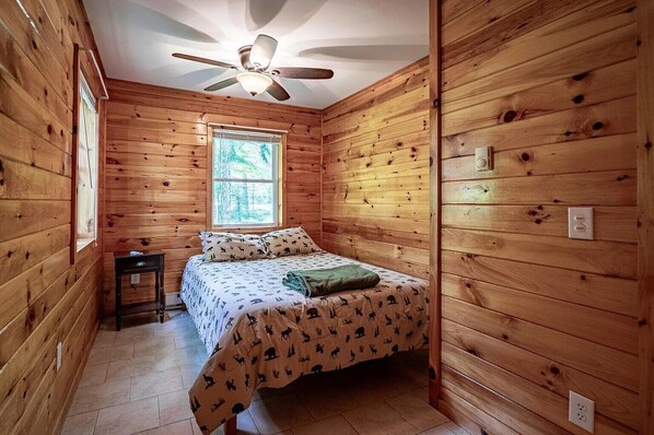 A wooden cabin room with a bed, a window, a ceiling fan, and a small table.