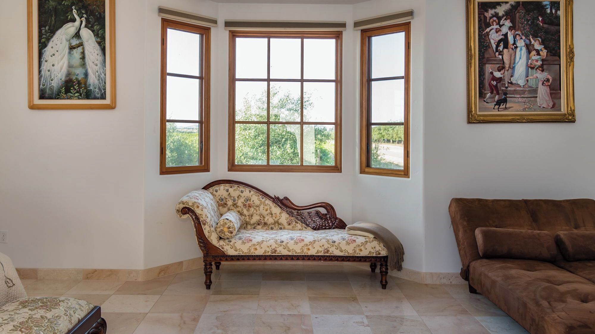 A room with a bay window, a vintage sofa, a brown couch, and two paintings on the walls.