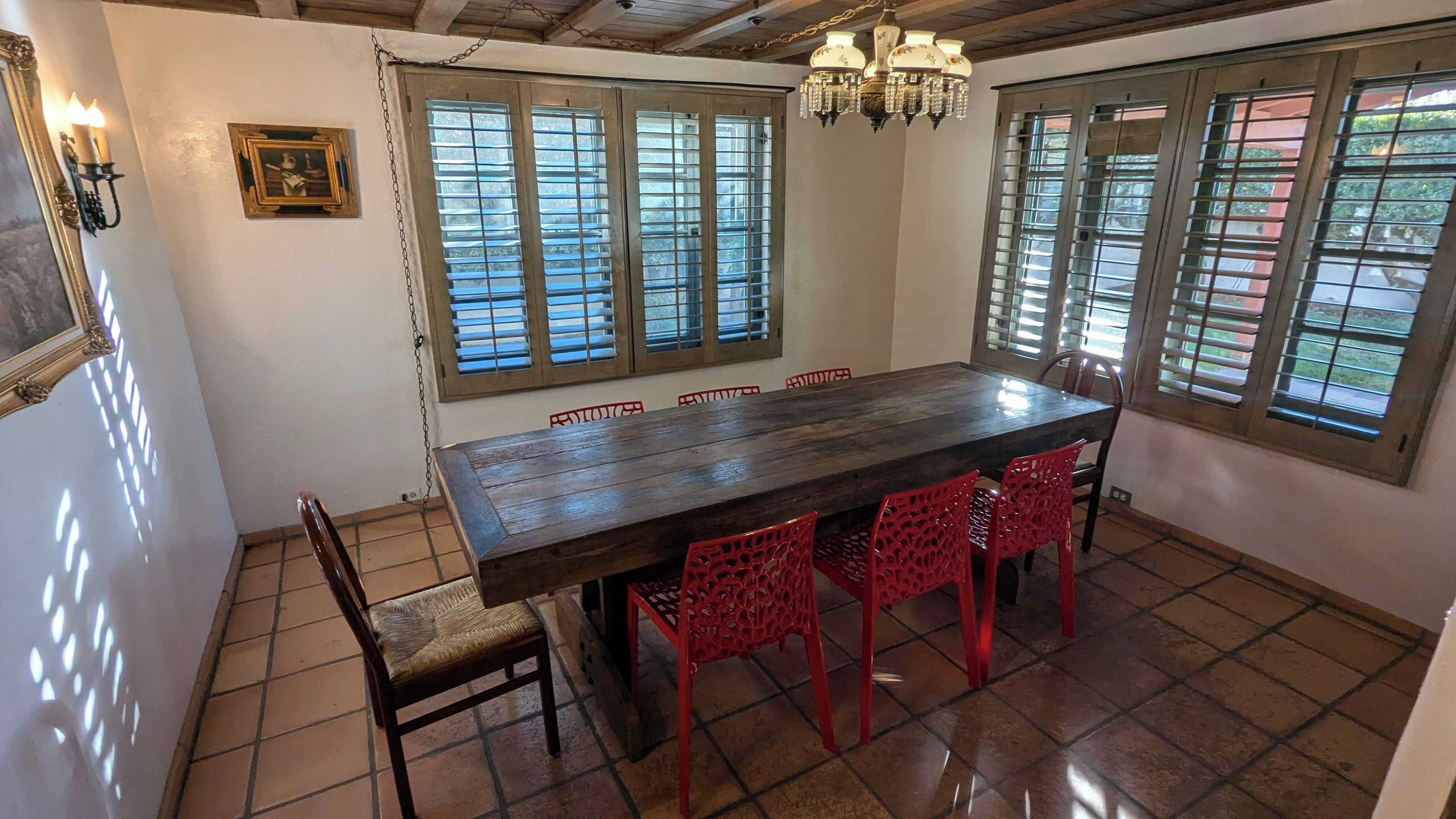 Un coin repas avec une table en bois et des chaises rouges, de grandes fenêtres équipées de stores et un lustre.