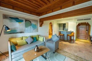 A living room with a sofa, armchair, and a coffee table. There is a painting on the wall and a kitchen area visible through an archway. - Le Pavillon Du Golf (Marrakech)