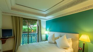 A hotel room with a bed, a television, a balcony with a view of greenery, and a lamp. - Le Pavillon Du Golf (Marrakech)