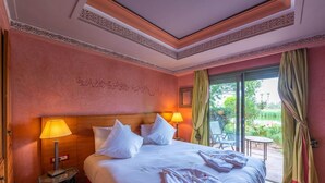 A hotel room with a large bed, a nightstand, a lamp, and a view of a garden through a glass door. - Le Pavillon Du Golf (Marrakech)