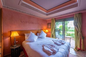 Quarto de hotel com uma cama grande, uma mesa de cabeceira, uma luminária e vista para um jardim através de uma porta de vidro.