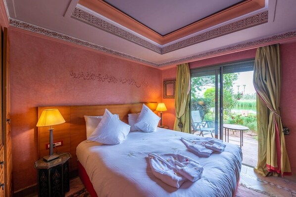 A hotel room with a large bed, a nightstand, a lamp, and a view of a garden through a glass door. - Le Pavillon Du Golf (Marrakech)