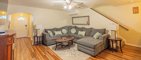 A living room with a sectional sofa, two side tables, a coffee table, hardwood flooring, a ceiling fan, and a staircase.