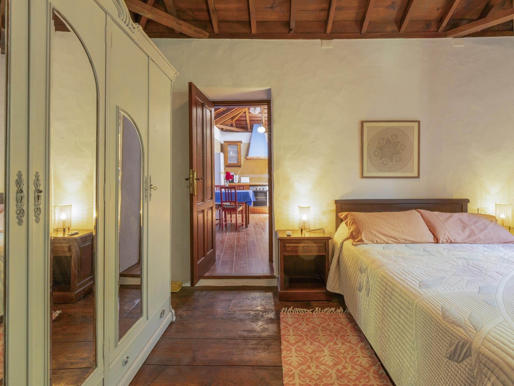 Quarto com teto de madeira, cama com almofadas, mesa de cabeceira, abajur e porta que dá para uma sala de jantar.