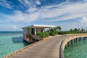 A wooden overwater structure with a covered seating area and a small building, surrounded by clear turquoise water and greenery.