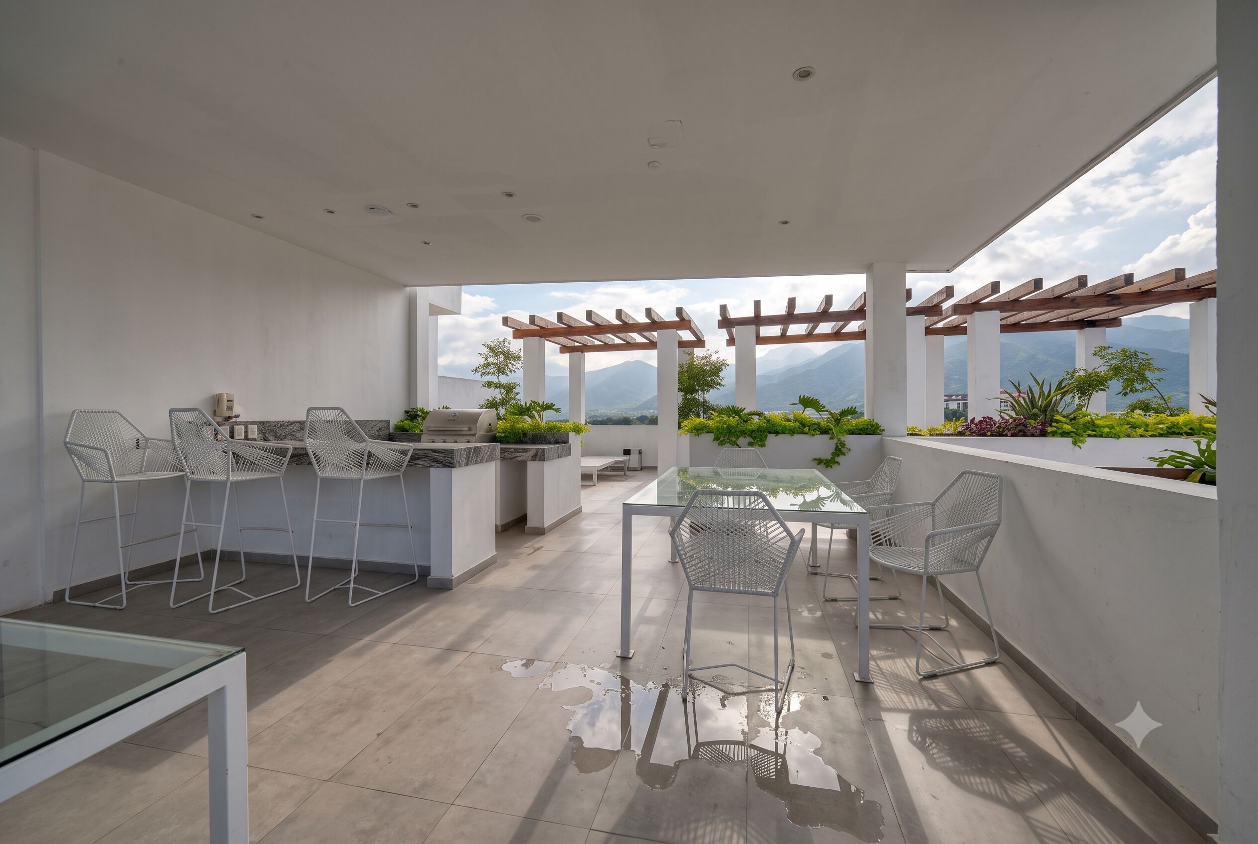 An outdoor patio with a glass table and chairs, a barbecue area, and a view of mountains.