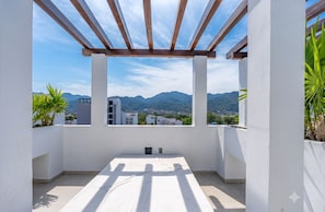 En balkon med udsigt til bjerge og bygninger, en træpergola og krukker med planter.