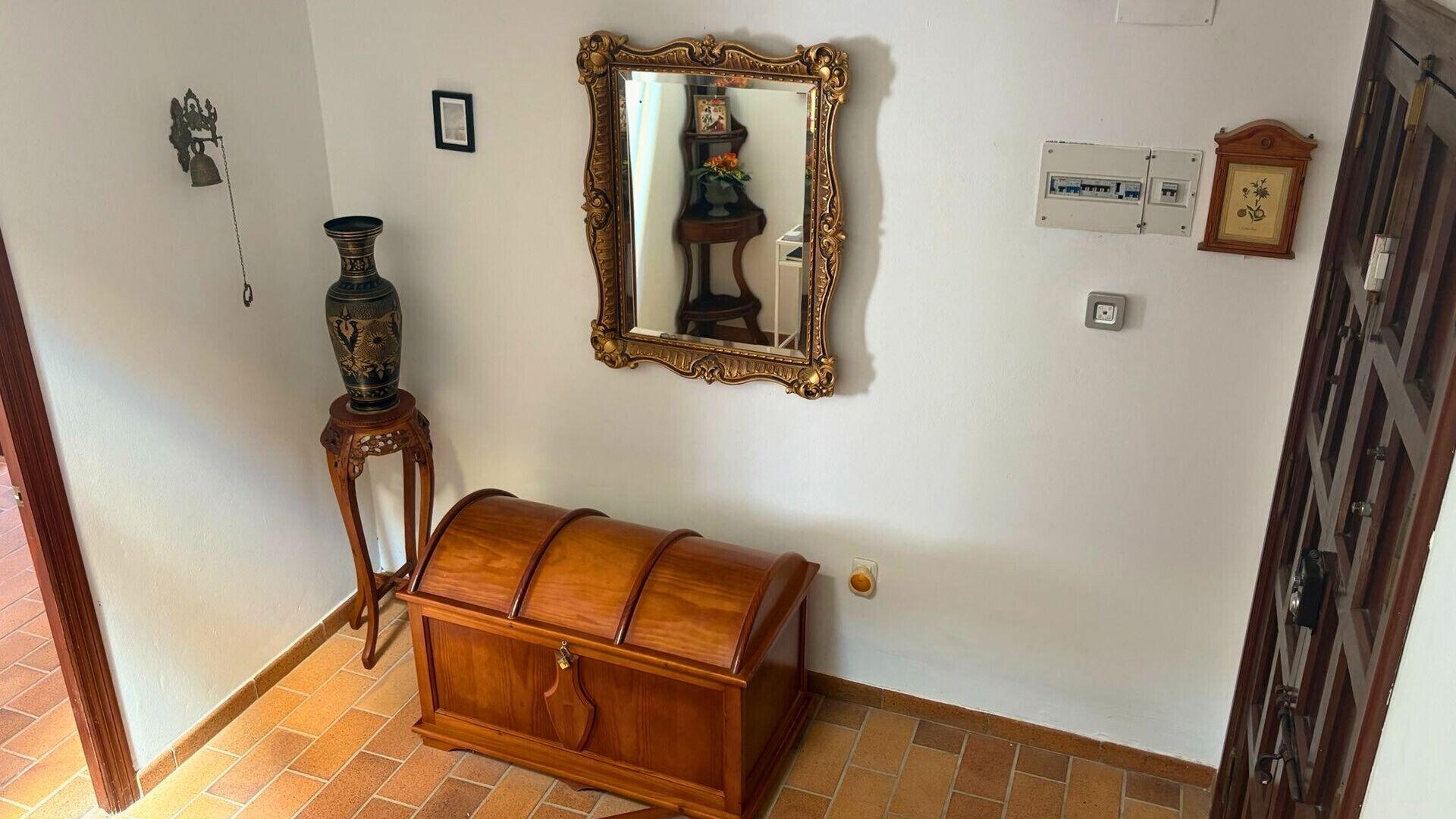 Une pièce comprenant un coffre en bois, un miroir fixé au mur, une petite table avec un vase et une horloge murale.